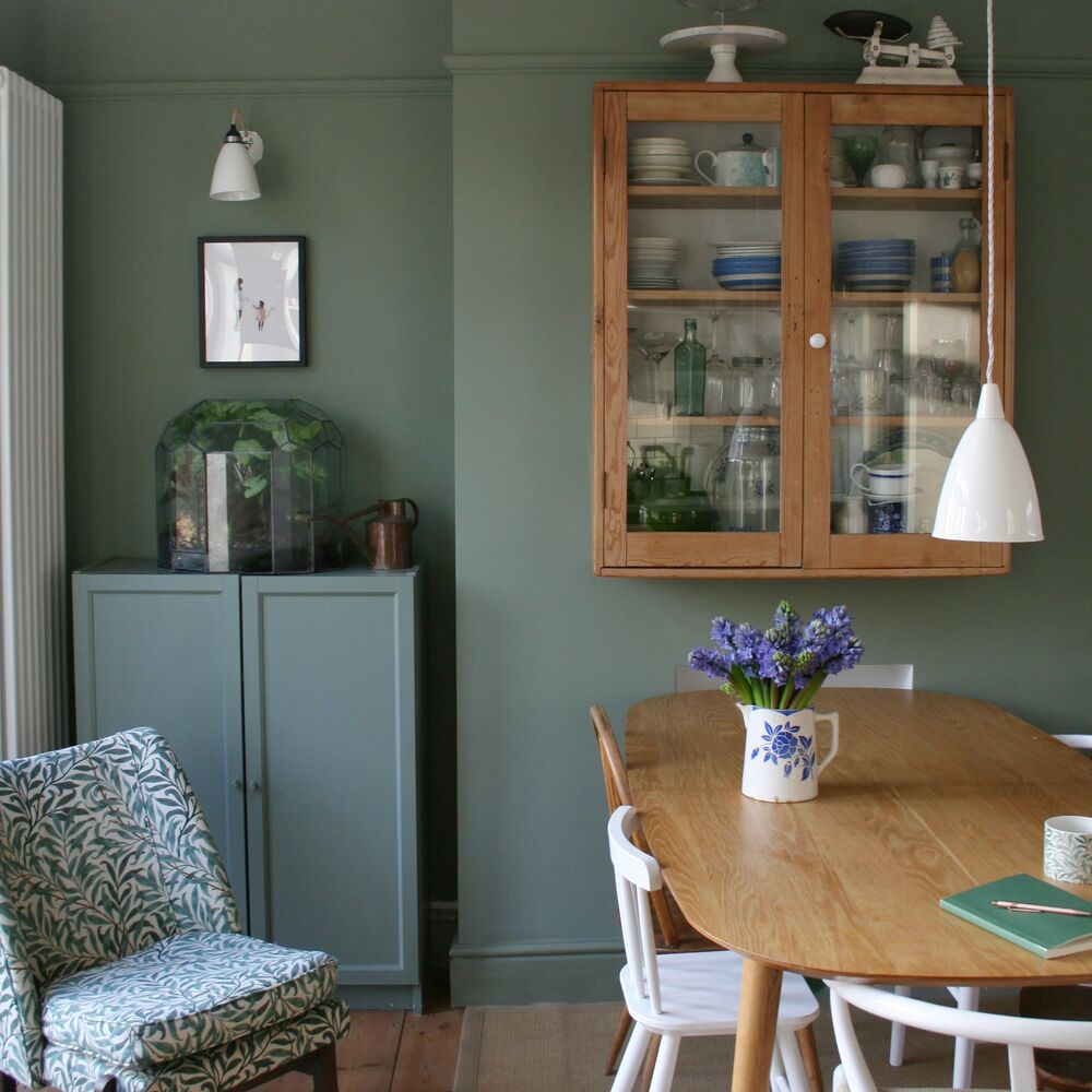A styled dining room corner featuring a light oak wooden hutch filled with dishes on a sage green wall, alongside a matching wooden dining table, white chairs, and a patterned armchair.