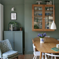 A styled dining room corner featuring a light oak wooden hutch filled with dishes on a sage green wall, alongside a matching wooden dining table, white chairs, and a patterned armchair.