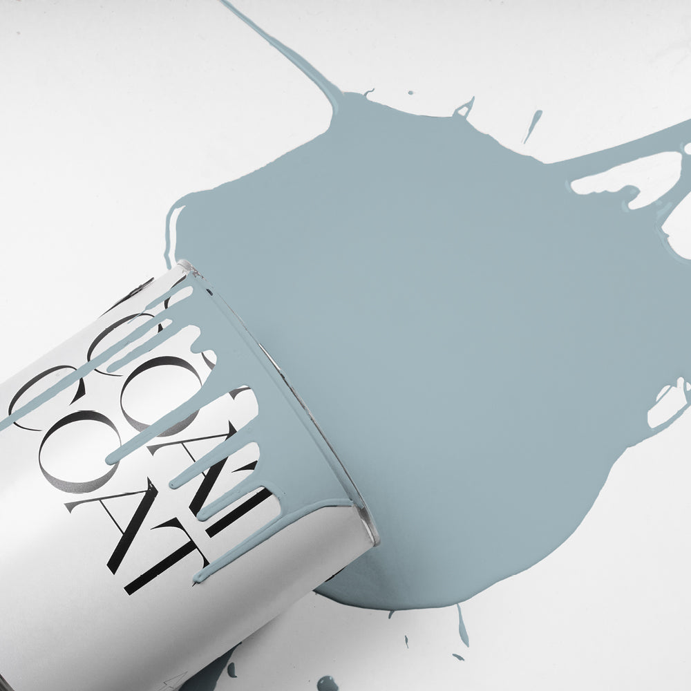 Paint splash showing Lie-in, a soft grey-blue colour by COAT Paints, poured from a tin